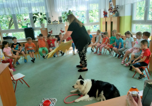 Dzieci siedzą na krzesełkach w dużym półkolu. Pani Beata rozkłada urodzinową matę na dywanie.