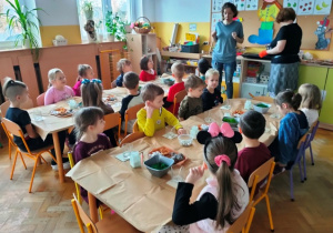 Dzieci siedzą przy stolikach, na których przygotowane są w pojemnikach materiały do pracy.