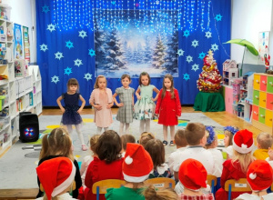 Dziewczynki śpiewają kolędę przed publicznością na tle świątecznej dekoracji.