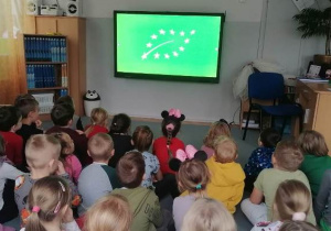 Dzieci siedzą na dywanie. Przygotowane są do projekcji edukacyjnej.