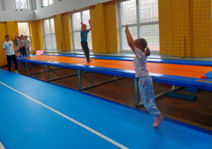 Dzieci poruszają się po trampolinie wykonując podskoki obunóż z rękoma uniesionymi do góry.