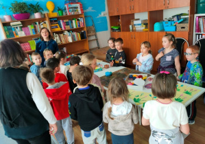 Dzieci w świetlicy stoją wokół stolików na których rozłożone są materiały plastyczne. Będą tworzyć własne zakładki do książki.