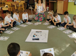 Dzieci siedzą na dywanie w kole i wykonują zadanie przygotowane przez muzealnika.