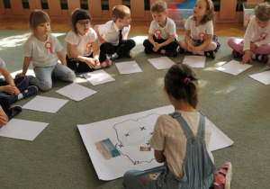 Dzieci siedzą na dywanie w kole. Poznają mapę Polski i zaznaczają na niej różne miasta.