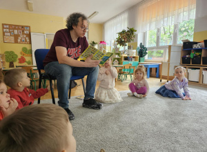 Mężczyzna siedzi na krześle i czyta książkę dzieciom siedzącym w kole.