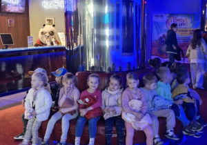 Dzieci siedzą na czerwonych kanapach i czekają na wejście do sali kinowej.