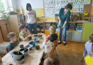 Dzieci obserwują panią Kasię, która pokazują jak należy wsypywać kolorowe grys do naczynia.