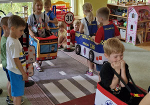 Dzieci poruszają się po ulicy jako uczestnicy ruchu drogowego, kierując różnymi pojazdami. Dwoje dzieci czeka na zielone światło stojąc przed pasami.