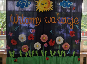 Dekoracja na tle której występowały dzieci - chmury, słońce, łąka z kwiatami i owadami oraz napis: Witamy wakacje.