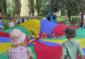 Dzieci trzymają chustę animacyjną i podrzucają nią kolorowe piłeczki.