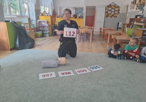 Dzieci siedzą na dywanie i przypominają sobie podstawowe numery alarmowe.