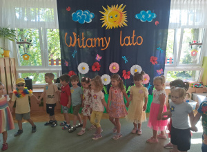 Dzieci trzymają się za ręce i stoją przed dekoracją - łąką i napisem: Witamy lato.