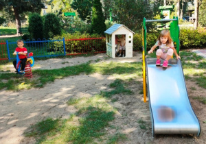 Dzieci korzystają ze sprzętu ogrodowego- bujaków, zjeżdżalni i domku.