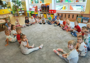 Dzieci siedzą na dywanie w dużym kole, słuchają wypowiedzi cioci na temat zdrowego jedzenia.