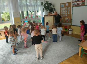 Dzieci stoją na dywanie w luźnej gromadce- naśladują ruch wykonywany przez instruktora.