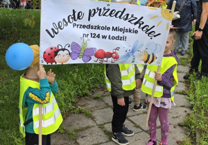 Dzieci trzymają transparent z napisem: Wesołe Przedszkole! Przedszkole Miejskie nr 124 w Łodzi.