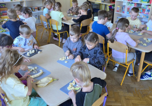 Dzieci siedzą przy stolikach i kroją owoce do sałatki.