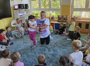 Dzieci siedzą na dywanie w kole. Tata Huga pokazuje ruchy taneczne wspólnie z jedną dziewczynką.