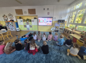 Dzieci siedzą na dywanie i oglądają bajkę edukacyjną na tablicy multimedialnej.