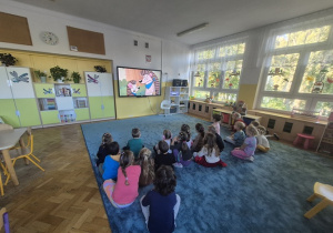 Dzieci siedzą na dywanie i oglądają bajkę edukacyjną.