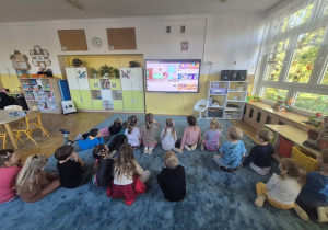 Dzieci siedzą na dywanie i oglądają bajkę edukacyjną.