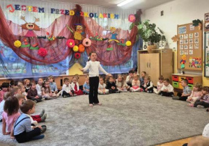 Dziewczynka z najstarszej grupy stojąc na środku dywanu śpiewa okolicznościową piosenkę.