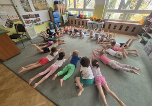 Dzieci leżą na dywanie i na sygnał nauczyciela podnoszą wyprostowane nogi i ręce.