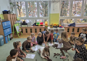 Dzieci siedzą na dywanie i układają za pomocą kasztanów wzory na kartkach.