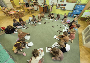 Dzieci siedzą na dywanie i układają kształty z kasztanów.