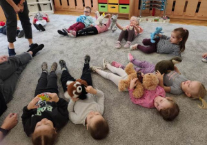 Dzieci leżą na dywanie na plecach z przytulanką położoną na brzuchu- oddychając unoszą maskotkę do góry.