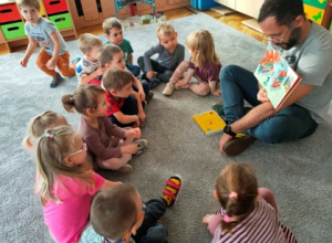 Dzieci skupione wokół prowadzącego oglądają ilustracje w książce.