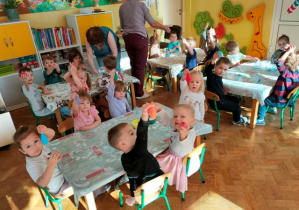 Dzieci siedząc przy stolikach podnoszą w górę rękę z wykonanym przez siebie gniotkiem.