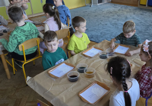 Dzieci siedzą przy stolikach i przyglądają się przedmiotom, które wykorzystają podczas warsztatów. Zastanawiają się do czego służą i co będzie tematem spotkania.