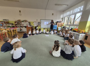 Dzieci siedzą na dywanie i oglądają prezentację multimedialna na tablicy.