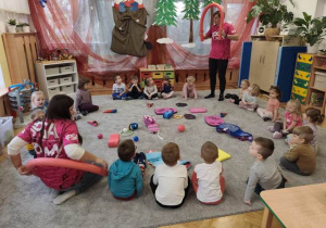 Dzieci siedzą w dużym kole na dywanie po środku którego rozłożone są akcesoria pływackie. Słuchają wypowiedzi trenera.