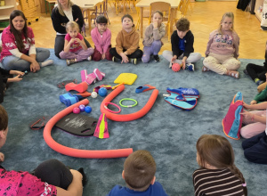 Dzieci siedzą na dywanie w kole i rozmawiają z instruktorem pływania. Na dywanie leżą przedmioty wykorzystywane podczas zajęć i zabaw w wodzie.
