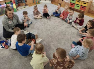 Dzieci siedzą na dywanie w dużym kole. Oglądają ilustracje w książce przeczytanej przez gościa.