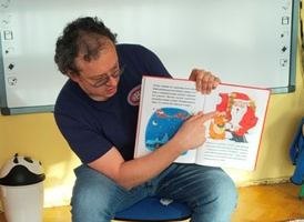 Mężczyzna siedzi na krześle, w ręku trzyma książkę dla dzieci.