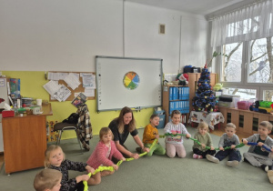 Dzieci siedzą na dywanie i bawią się gumą sensoryczną, ugniatając ją i rozciągając w różnych kształtach.