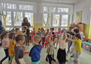 Dzieci tańczą na dywanie, trzymając gumę sensoryczną i poruszając się w rytm muzyki.