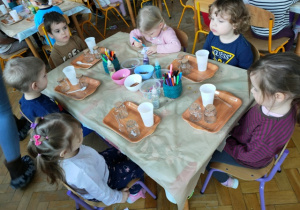 Dzieci czekają na dalsze wskazówki dotyczące pracy- przed nimi stoją tace na których ustawione są puste szklane pojemniki, oraz kubeczek z wodą.