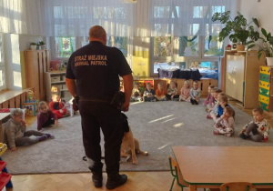 Dzieci siedzą w dużym półkolu na dywanie, uważnie słuchają wypowiedzi gościa.