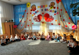 Dzieci siedzą na dywanie w dużym półkolu.