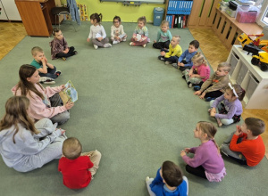 Dzieci siedzą na dywanie i słuchają czytanych bajek.