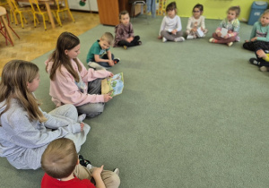 Dzieci siedzą na dywanie i zaciekawieniem słuchają czytanej bajki..