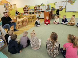 Dzieci siedzą na dywanie i zaciekawieniem słuchają opowiadania czytanego przez Pana Mariusza.