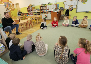 Dzieci siedzą na dywanie a Pan Mariusz czyta opowiadanie.