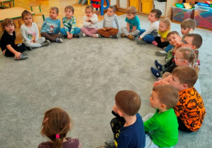 Dzieci siedzą w dużym półkolu na dywanie, czekając na przybycie gościa.
