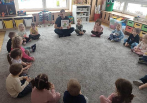 Dzieci siedzą w dużym kole na dywanie. Gość prezentuje dzieciom książkę, która będzie za chwilę czytana.