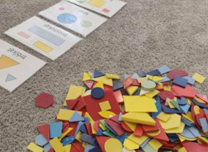 Na dywanie położone są karty edukacyjne, oraz plastikowe figury geometryczne.